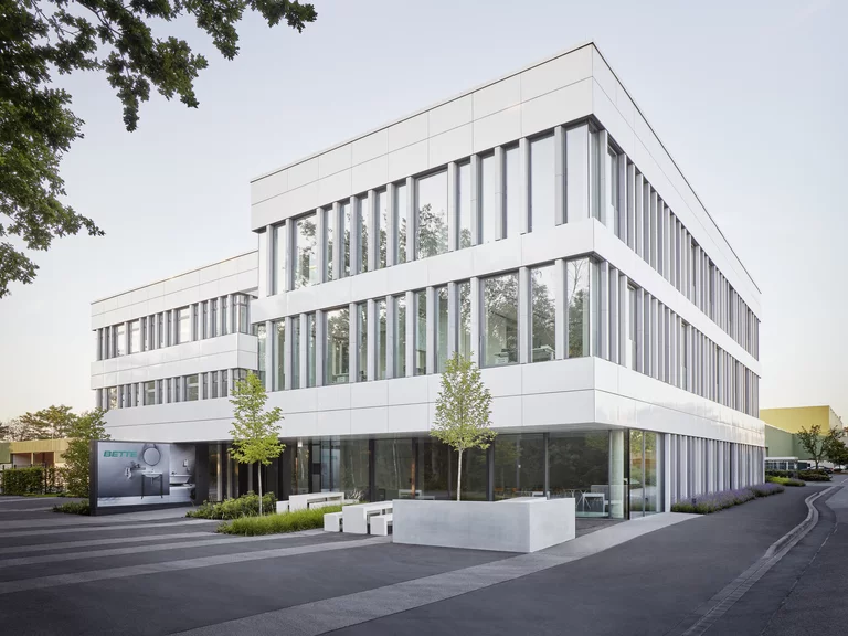 Verwaltungsgebäude von Bette mit klarer, geradliniger Architektur und großen Glasfassaden, umgeben von einer gepflegten Außenanlage mit Bäumen und Sitzmöglichkeiten.