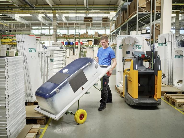 Ein Auszubildender zur Fachkraft für Lagerlogistik bei Bette transportiert eine verpackte Badewanne im Lager mit Hilfe eines Transportwagens.