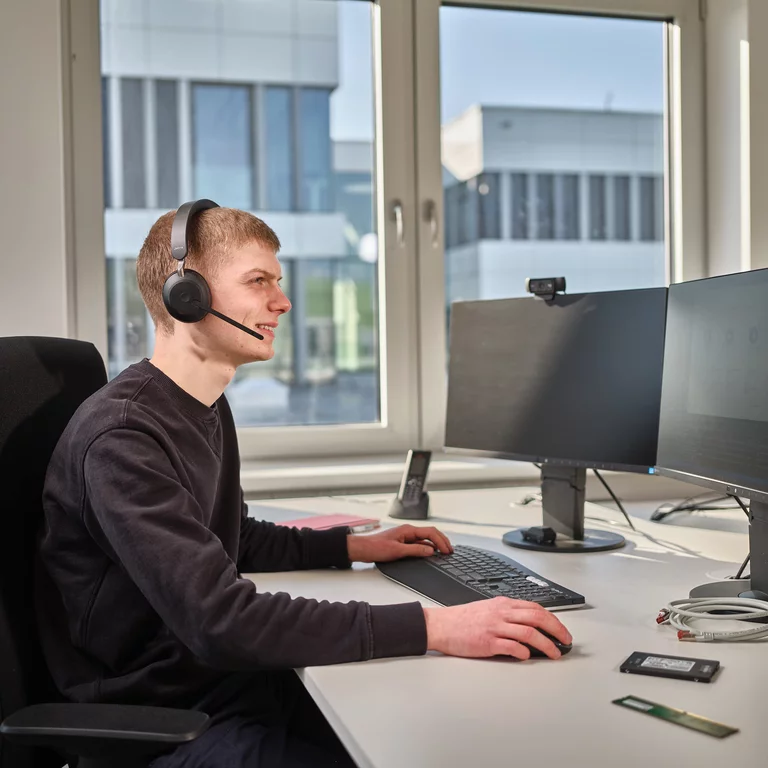 Mitarbeiter bei Bette sitzt mit Headset an einem Schreibtisch und arbeitet an einem dualen Monitor-Setup.