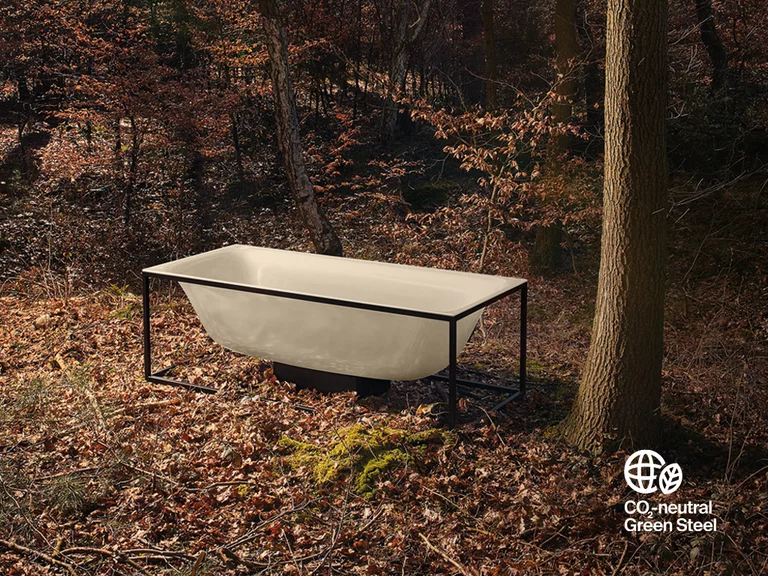 BetteLux Shape: Freistehende Bette Badewanne aus im herbstlichen Wald platziert. Unten rechts befindet sich ein weißes Logo mit Weltkugel und Blatt, das die CO₂-Neutralität des verwendeten Stahls hervorhebt.