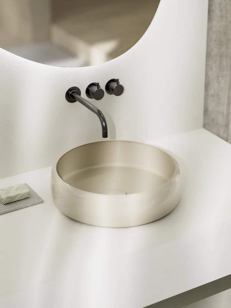 Beige, rond opzetwastafel met zwarte wandkraan, voor een organisch gevormde spiegel op een wit blad.