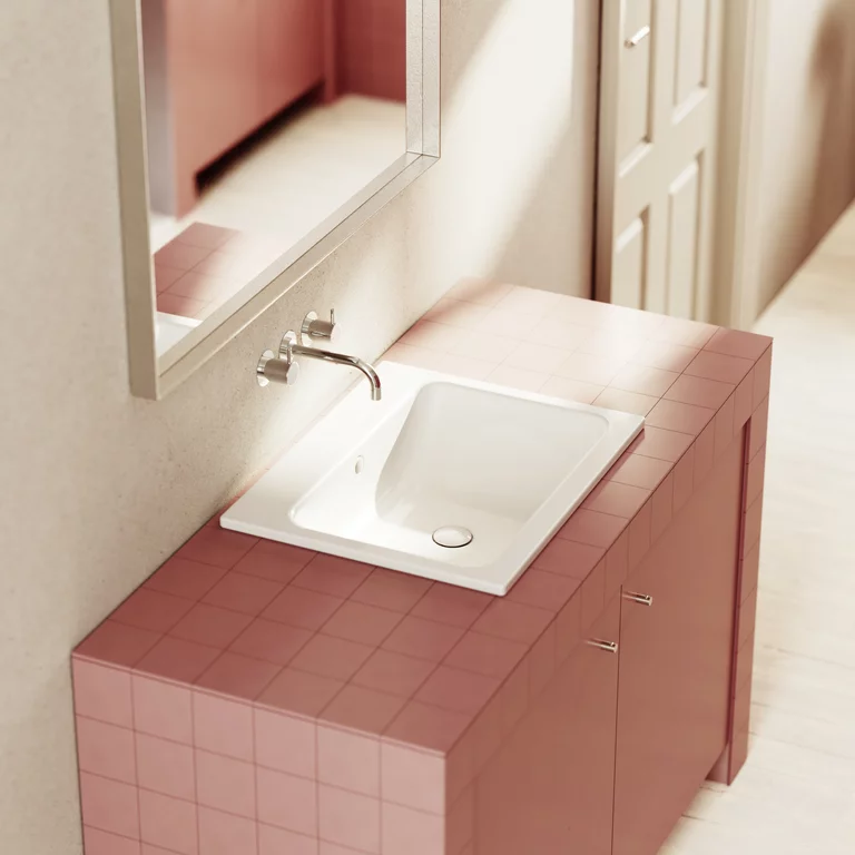 Waschtisch mit weißem Einbauwaschbecken, Wandarmatur und rosa Fliesen in einem hellen Badezimmer mit Spiegel.