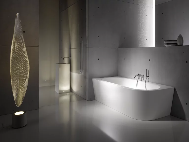 BetteStarlet IV Silhouette, una elegante bañera exenta de color blanco, colocada en un cuarto de baño minimalista con paredes de hormigón, iluminación ambiental y una moderna lámpara de pie.