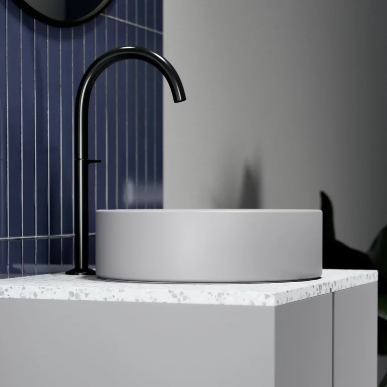 Vasque à poser BetteSuno avec vasque ronde de couleur silver sur un plan de travail en terrazzo clair, complété par une robinetterie noire, devant un mur élégant avec des carreaux bleus verticaux et une décoration végétale discrète en arrière-plan.