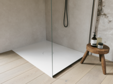 Witte inloopdouchebak met glazen wand en zwarte douche, naast een klein houten krukje met verzorgingsproducten.