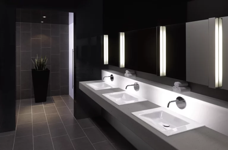 Три прямоугольные стальные эмалированные раковины на столешнице, современная ванная с зеркалом и подсветкой, тёмная плитка.