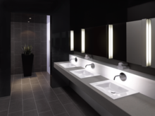 Три прямоугольные стальные эмалированные раковины на столешнице, современная ванная с зеркалом и подсветкой, тёмная плитка.