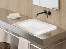 Современная квадратная раковина белого цвета на металлической консоли с чёрным настенным смесителем и полотенцами.