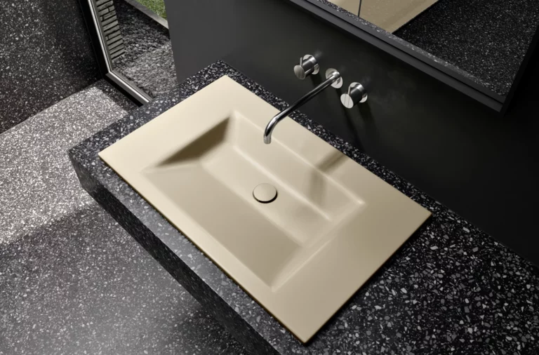 Прямоугольная стальная эмалированная раковина с четкими краями на тёмной столешнице, настенный смеситель сверху.