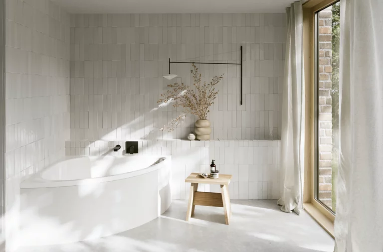 Helles Bad mit weißer Badewanne, großem Fenster, gefliesten Wänden, Hocker und sanftem Sonnenlicht auf dem Boden.