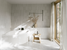 Helles Bad mit weißer Badewanne, großem Fenster, gefliesten Wänden, Hocker und sanftem Sonnenlicht auf dem Boden.
