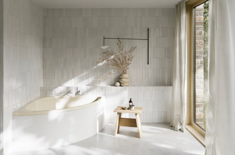 Helles Bad mit beiger Badewanne, bodentiefem Fenster, Vorhang, Hocker und Vase im sanften Sonnenlicht.
