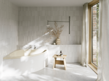 Helles Bad mit beiger Badewanne, bodentiefem Fenster, Vorhang, Hocker und Vase im sanften Sonnenlicht.