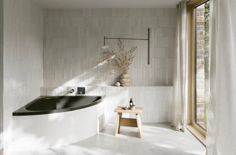 Helles Bad mit Badewanne mit dunkler Innenfläche, bodentiefem Fenster, Hocker und Vase im Sonnenlicht.