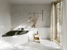 Helles Bad mit Badewanne mit dunkler Innenfläche, bodentiefem Fenster, Hocker und Vase im Sonnenlicht.