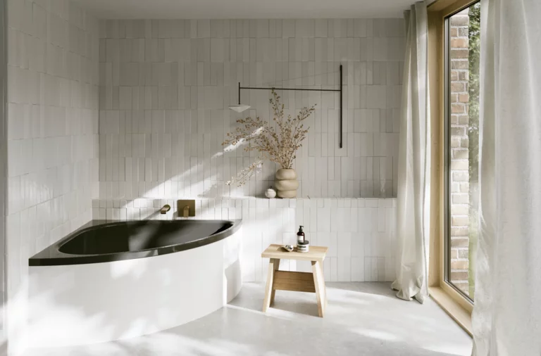 Helles Bad mit Badewanne mit dunkler Innenfläche, bodentiefem Fenster, Hocker und Vase im sanften Sonnenlicht.