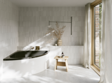 Helles Bad mit Badewanne mit dunkler Innenfläche, bodentiefem Fenster, Hocker und Vase im sanften Sonnenlicht.