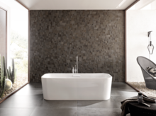 Отдельностоящая прямоугольная ванна из стальной эмали перед фактурной тёмной стеной в современной ванной.