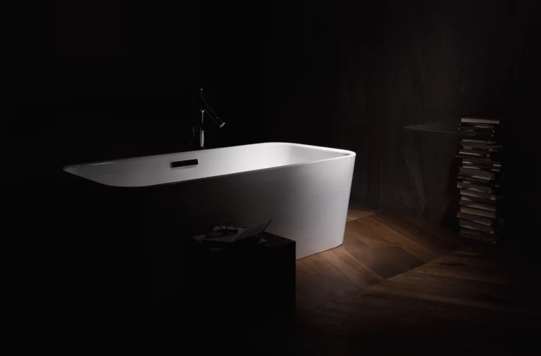 Отдельно стоящая прямоугольная ванна из стальной эмали на деревянном полу, рядом стопка книг и небольшой столик.