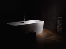 Отдельно стоящая прямоугольная ванна из стальной эмали на деревянном полу, рядом стопка книг и небольшой столик.