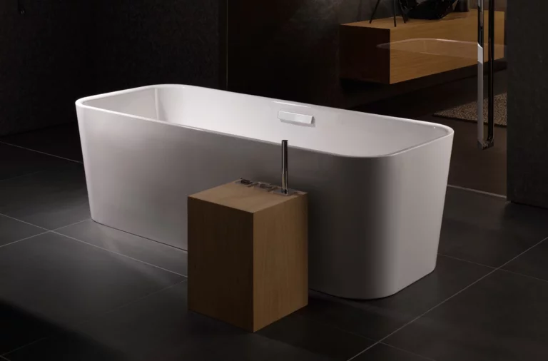 Freistehende, rechteckige Badewanne aus Stahl-Email, weiß, mit modernem Holzelement und Armatur auf dunklem Fliesenboden.