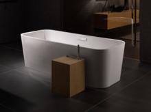 Отдельностоящая прямоугольная ванна из стальной эмали, белая, с современным деревянным элементом и смесителем на тёмном кафельном полу.