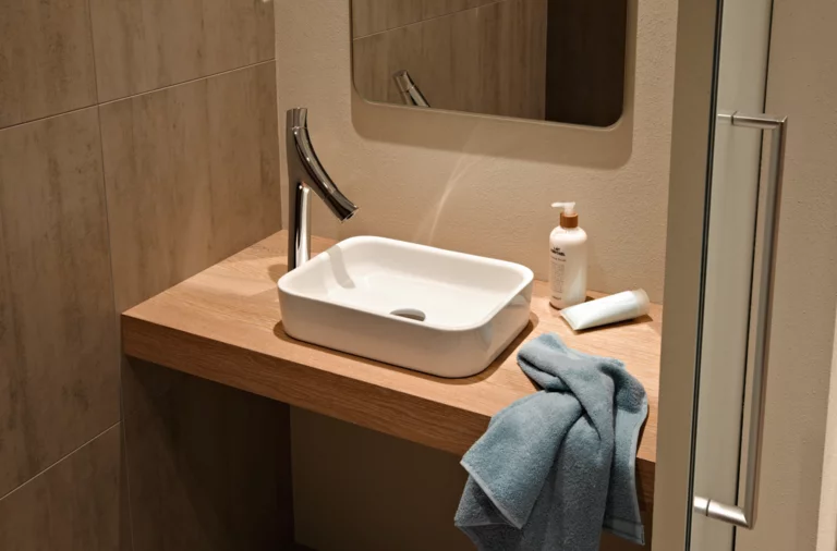 Rechteckiges Stahl-Email-Waschbecken auf Einbauplatte, moderner Wasserhahn, Spiegel, Handtuch und Pflegeprodukte daneben.