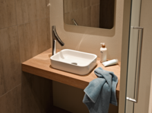 Rechteckiges Stahl-Email-Waschbecken auf Einbauplatte, moderner Wasserhahn, Spiegel, Handtuch und Pflegeprodukte daneben.