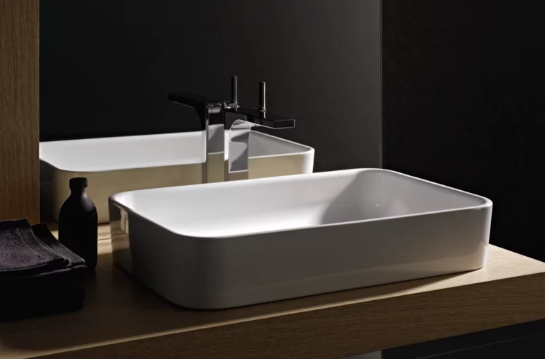 Lavabo rectangulaire en acier émaillé sur plan clair avec robinet, à côté serviette foncée et petite bouteille.