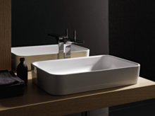 Lavabo rectangulaire en acier émaillé sur plan clair avec robinet, à côté serviette foncée et petite bouteille.