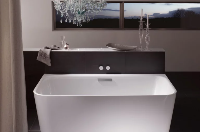 Freestanding rectangular steel-enamel bathtub in modern bathroom with chandelier and window in background.