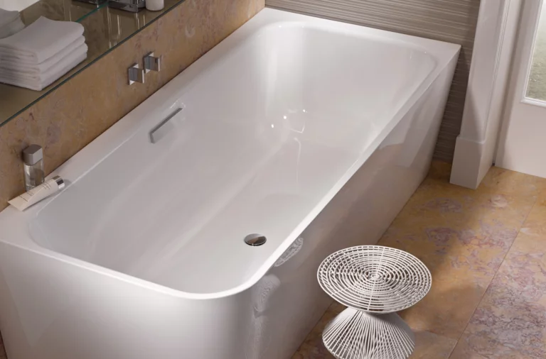 Rectangular steel-enamel bathtub, glossy white, with shelf and modern stool next to it in bathroom.