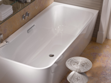 Rectangular steel-enamel bathtub, glossy white, with shelf and modern stool next to it in bathroom.