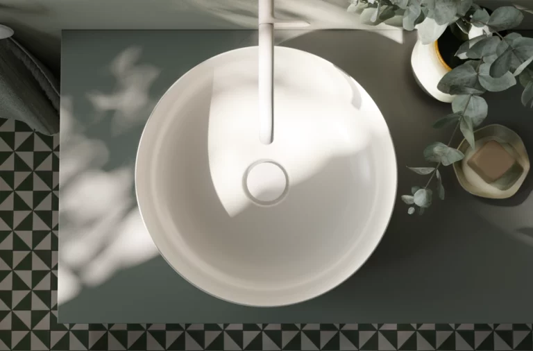 Rundes Waschbecken aus Stahl-Email auf Einbauplatte, weiße Armatur, dekorative Pflanzen, geometrische Fliesen am Boden.