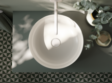Rundes Waschbecken aus Stahl-Email auf Einbauplatte, weiße Armatur, dekorative Pflanzen, geometrische Fliesen am Boden.