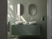 Zwei runde Waschbecken auf grüner Einbauplatte, zwei Spiegel, Stehlampe, modernes Bad mit geometrischem Fliesenboden.