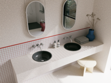 Helles Badezimmer mit zwei dunkelgrauen Waschtischen, ovalen Spiegeln, gefliester Ablage und roter Linie an der Wand.