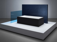 Rechteckige Badewanne aus Stahl-Email mit schwarzer Verkleidung, auf erhöhtem Podest, vor blauer Wand und Glasabtrennung.