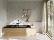 Helles Bad mit rechteckiger Badewanne mit dunkler Innenfläche, beiger Umrandung, bodentiefem Fenster und Vorhang.