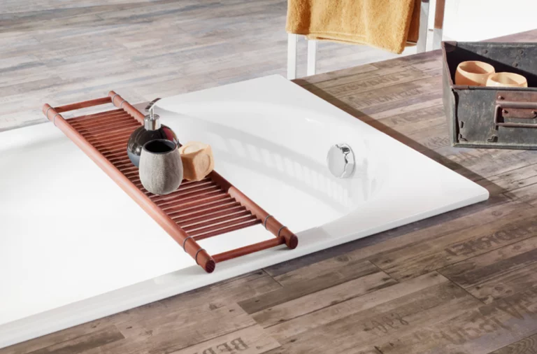 Rectangular steel enamel bathtub, flat built-in, with wooden tray, soap dispenser and soap, on wooden floor.