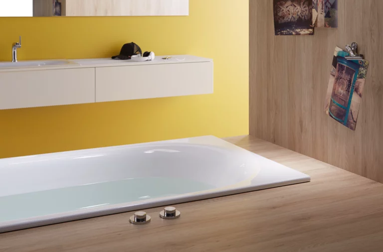 Rectangular steel-enamel bathtub, partially set into wooden floor, countertop with washbasin against yellow wall.