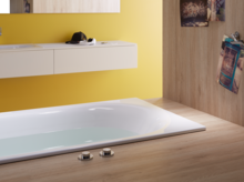 Rectangular steel-enamel bathtub, partially set into wooden floor, countertop with washbasin against yellow wall.