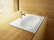 Ovales Stahl-Email Waschbecken auf Einbauplatte, mit Einzelarmatur, umgeben von heller Holzoberfläche.