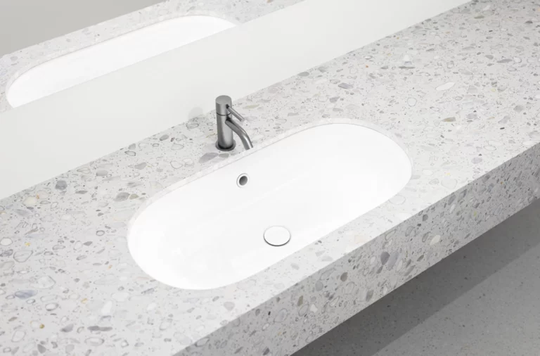 Oval white inset washbasin in terrazzo-style countertop with silver faucet and mirror in the background.
