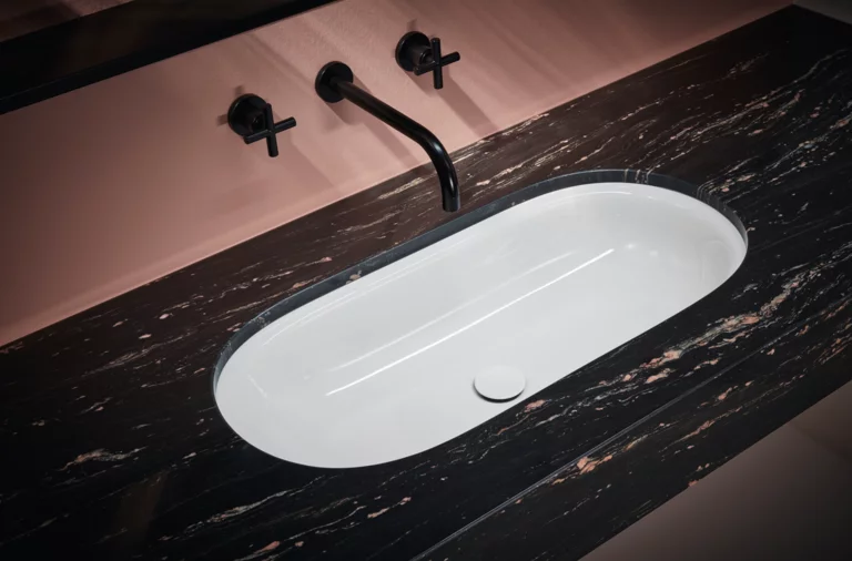 Lavabo da incasso ovale bianco in piano in marmo scuro con rubinetto nero a parete e sfondo rosa.