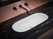 Oval white inset washbasin in dark marble countertop with black wall-mounted faucet and pink wall background.