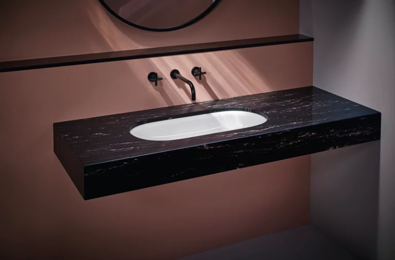 Oval white inset washbasin in a floating dark marble console with black wall-mounted faucet and pink wall background.