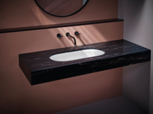 Oval white inset washbasin in a floating dark marble console with black wall-mounted faucet and pink wall background.