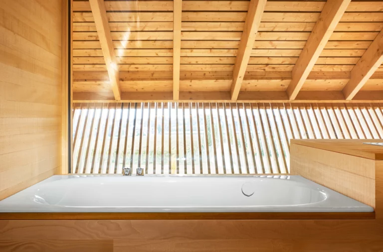 Baignoire rectangulaire en acier émaillé, encastrée dans un habillage bois, devant grande fenêtre à lamelles en bois.