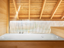 Baignoire rectangulaire en acier émaillé, encastrée dans un habillage bois, devant grande fenêtre à lamelles en bois.